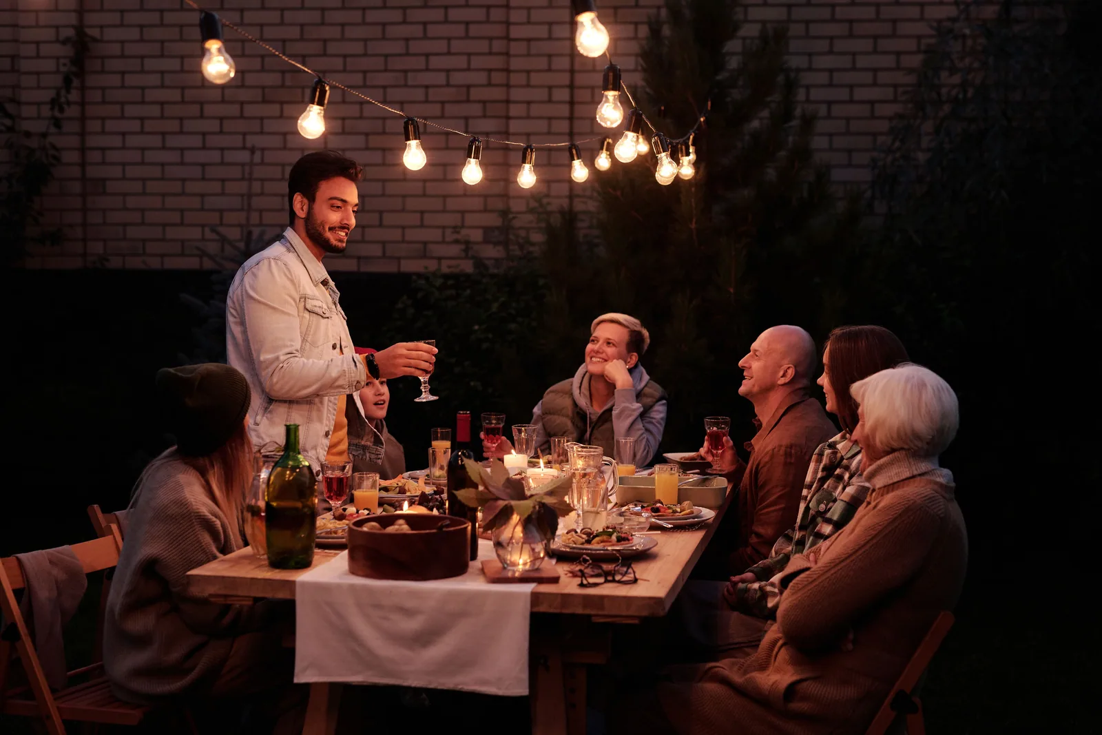Guests seated around a long backyard table lit by hanging string lights after dark