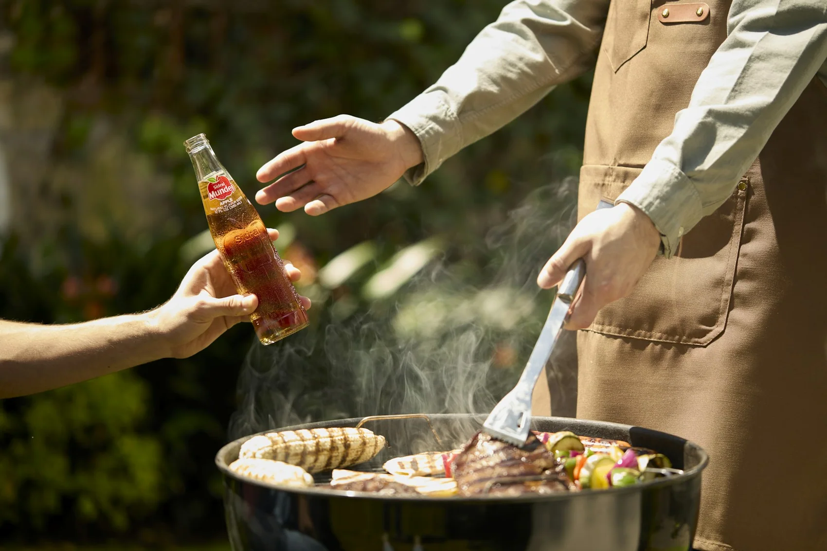 A cook turning food on the grill while someone passes across a cold drink