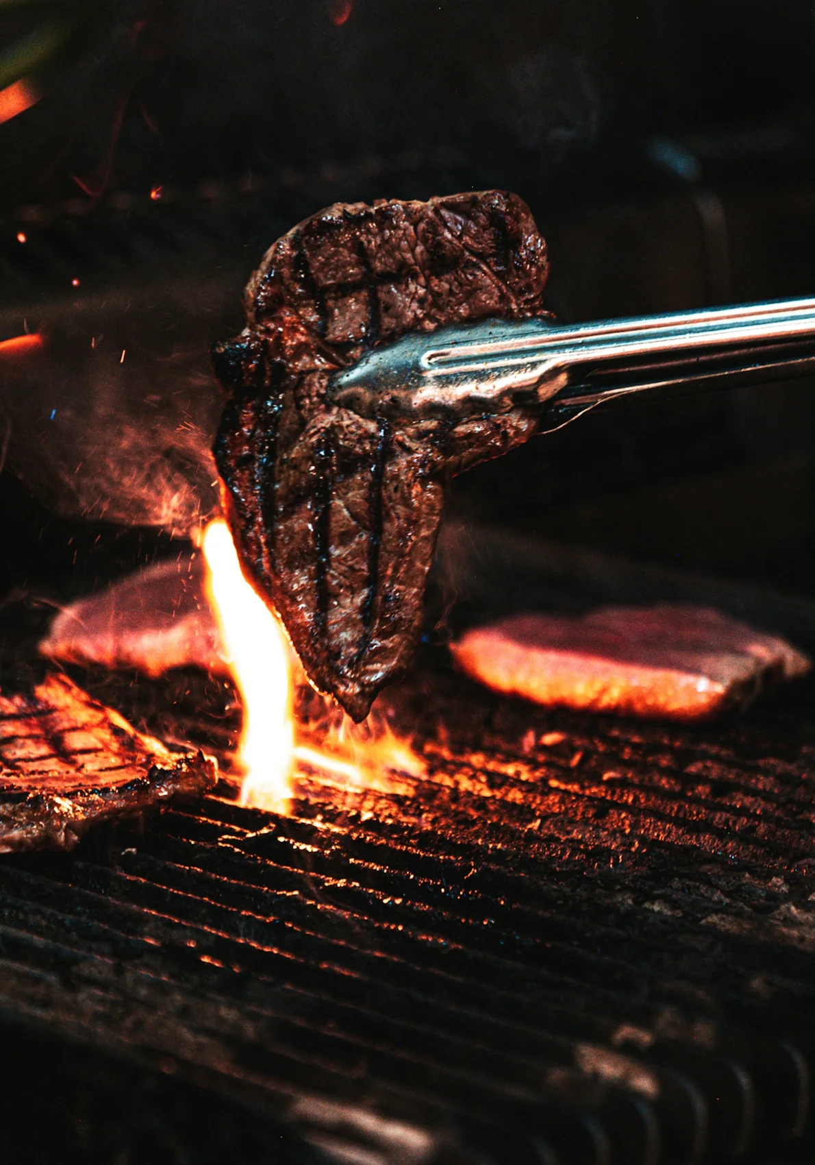 Tongs lifting a seared steak clear of the flames