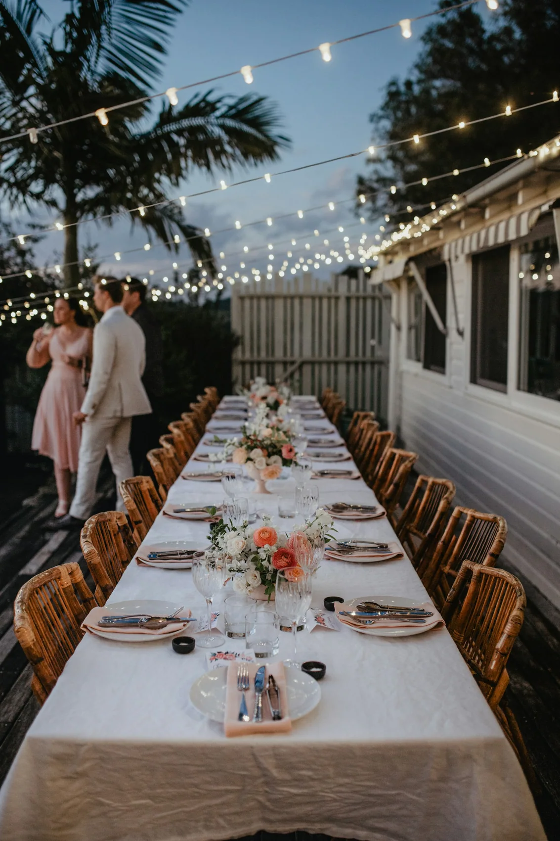 A long banquet table set end to end outdoors beneath hanging lights, before guests arrive